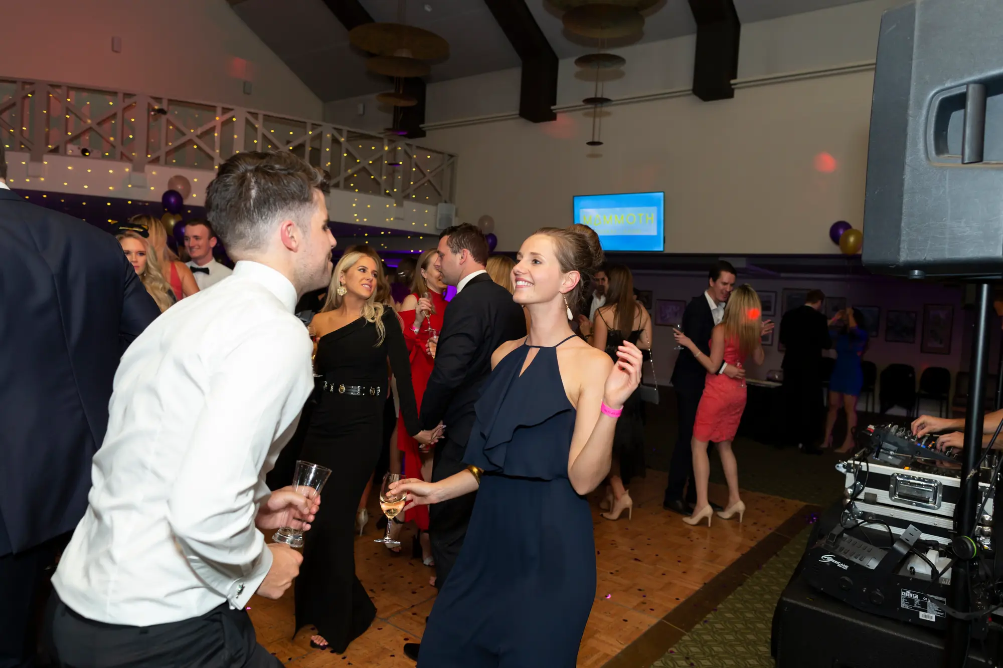 kooyong lawn tennis club masquerade ball navy blue dance floor couple A lively, high-energy moment on the dance floor at the Kooyong Lawn Tennis Club End-of-Year Masquerade Ball, featuring a man in a white shirt and black trousers smiling and holding a glass while interacting with a woman in a navy blue ruffled halter-neck dress with a pink wristband. Guests in tuxedos and cocktail dresses dance under vibrant purple lighting, with fairy lights on the mezzanine, a DJ booth in the corner, and clusters of purple and gold balloons adding festive flair. The image captures joyful celebration,