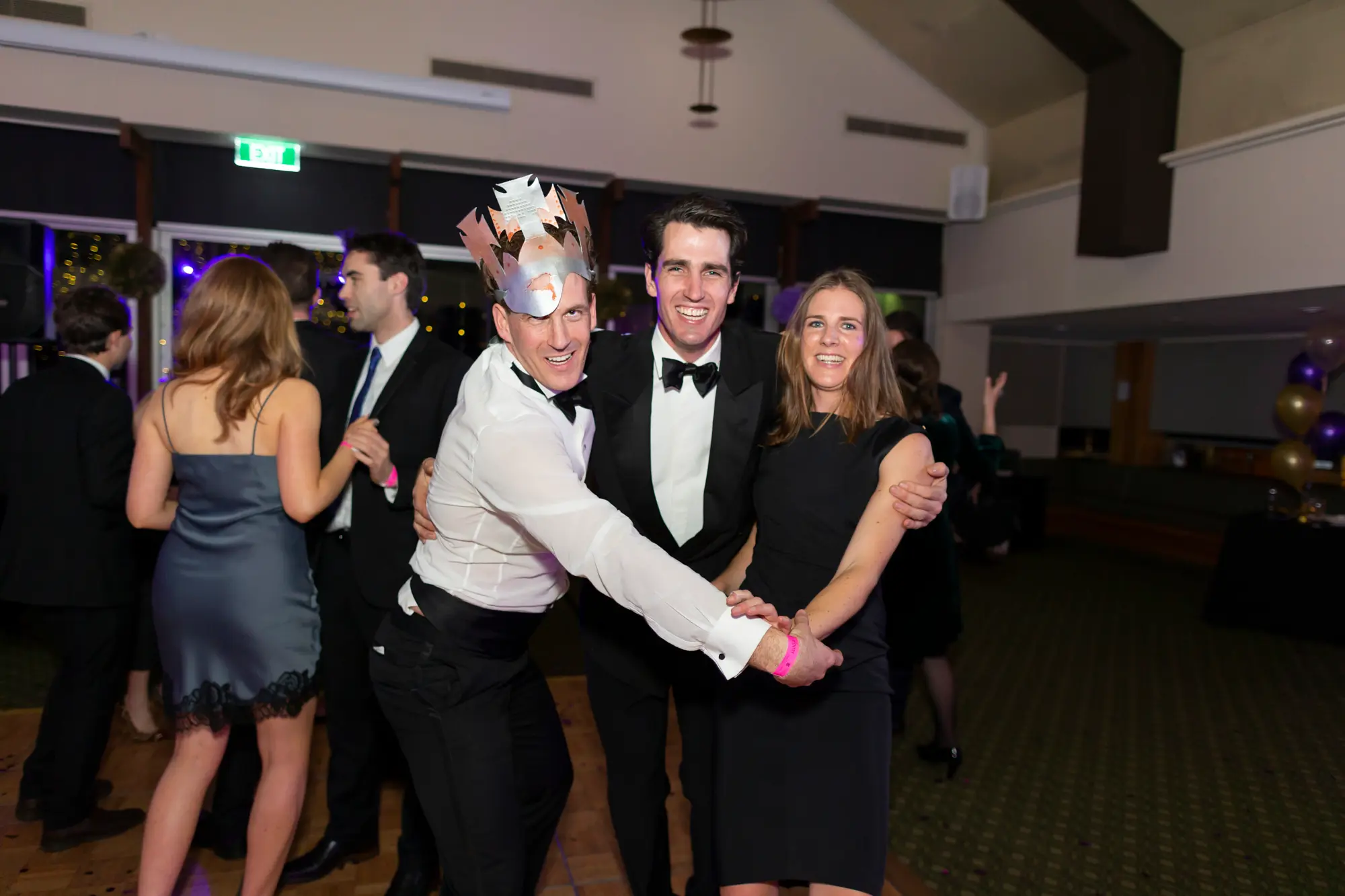 kooyong lawn tennis club masquerade ball paper crown black Three guests pose together at the Kooyong Lawn Tennis Club End-of-Year Masquerade Ball in Toorak. A man on the left wears a white shirt, black trousers, and a festive multi-colored paper crown, crouching with a wide smile. In the center, a man in a black tuxedo and bow tie smiles warmly with his arm around a woman in a black cocktail dress on the right. Pink event wristbands are visible on all three, and clusters of purple and gold balloons, wood paneling, and mingling guests are seen in the festive indoor background.