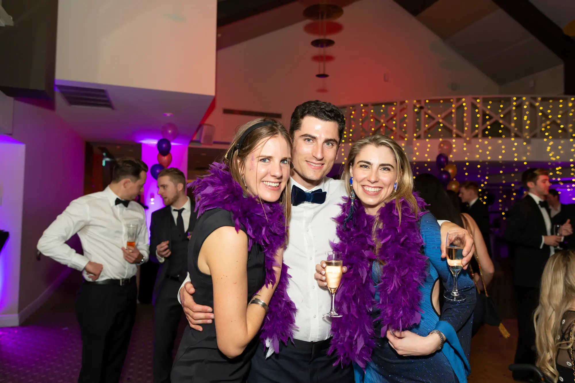 kooyong lawn tennis club masquerade ball purple feather boa group champagne Three guests pose closely together wearing purple feather boas at the Kooyong Lawn Tennis Club End-of-Year Masquerade Ball, smiling and holding champagne as they enjoy the lively celebration. Surrounded by fairy lights, purple and gold balloons, and elegantly dressed guests, the image captures the energetic and social atmosphere of the black-tie year-end event.
