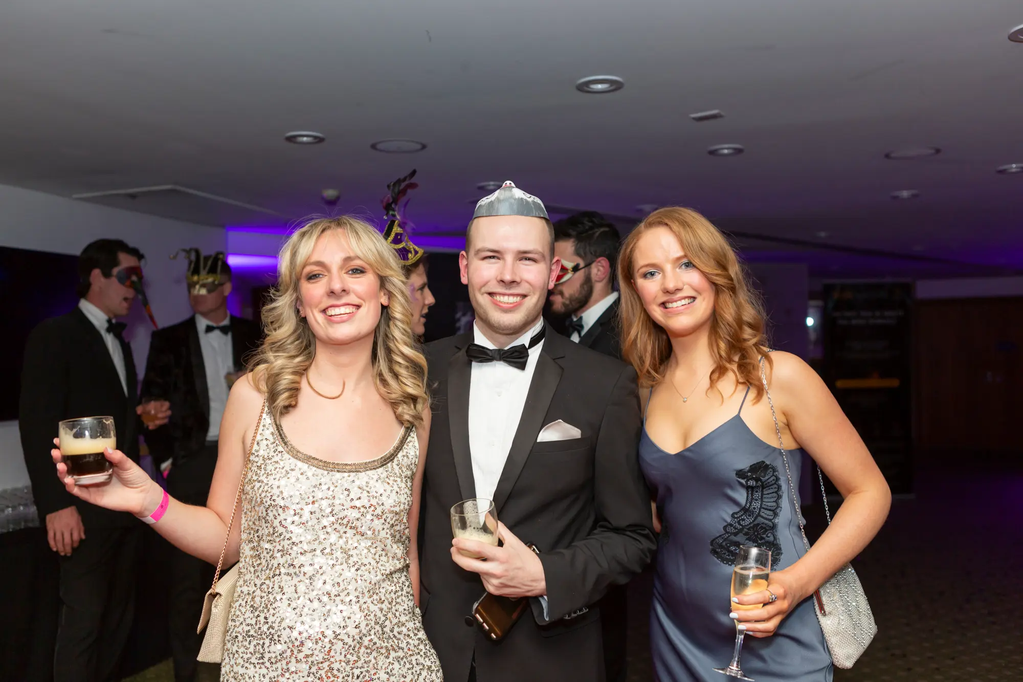 kooyong lawn tennis club masquerade ball three guests posing with drinks Three guests pose together at the Kooyong Lawn Tennis Club End-of-Year Masquerade Ball in Toorak. A woman on the left wears a silver sequined dress and holds a dark drink, smiling broadly. The man in the center wears a black tuxedo, bow tie, and a silver masquerade mask or hat, holding a glass of champagne. A red-haired woman on the right wears a slate-blue slip dress with black lace detailing and holds a champagne flute. The indoor venue features ambient purple lighting, a bar or catering area in the background, and other formally dressed guests, creating an elegant and festive atmosphere.