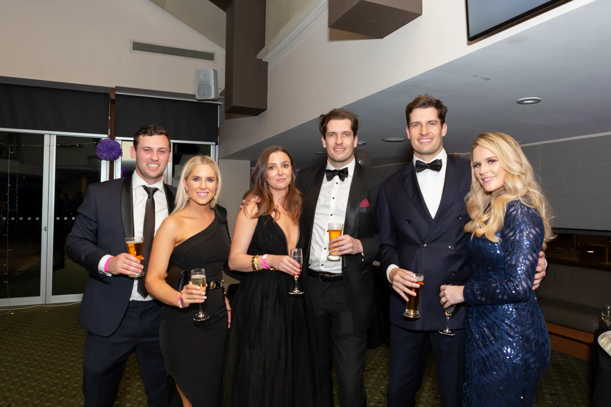 kooyong lawn tennis club masquerade ball yarrayong ball group portrait black wall six guests Six guests pose together at the Kooyong Lawn Tennis Club End-of-Year Masquerade Ball, also known as the YarraYong Ball, in Toorak. They are standing in front of a black wall and glass partition with a white floral display visible behind it. The group is dressed in formal evening attire, including black dresses, a navy blue sequined dress, and tuxedos, with some wearing masquerade masks. They are holding glasses of beverages, smiling, and enjoying a lively and celebratory indoor black-tie event atmosphere.