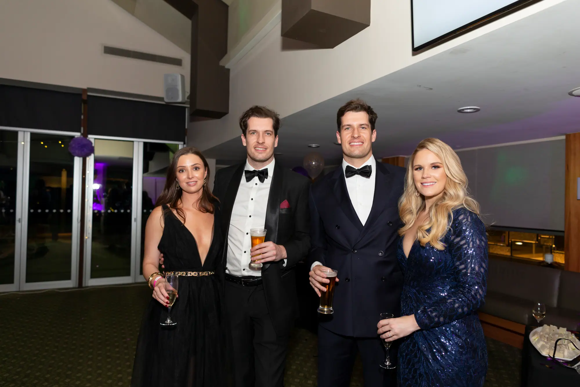 kooyong lawn tennis club masquerade ball yarrayong ball group portrait black wall Four guests pose together at the Kooyong Lawn Tennis Club End-of-Year Masquerade Ball, also known as the YarraYong Ball, in Toorak. They are standing in front of a black wall and glass partition with a glimpse of a white floral display behind it. All are dressed in formal evening attire—two women in elegant dresses and two men in black and navy tuxedos—holding glasses of champagne and beer. Warm ambient lighting highlights the celebratory and sophisticated atmosphere of this indoor black-tie event.