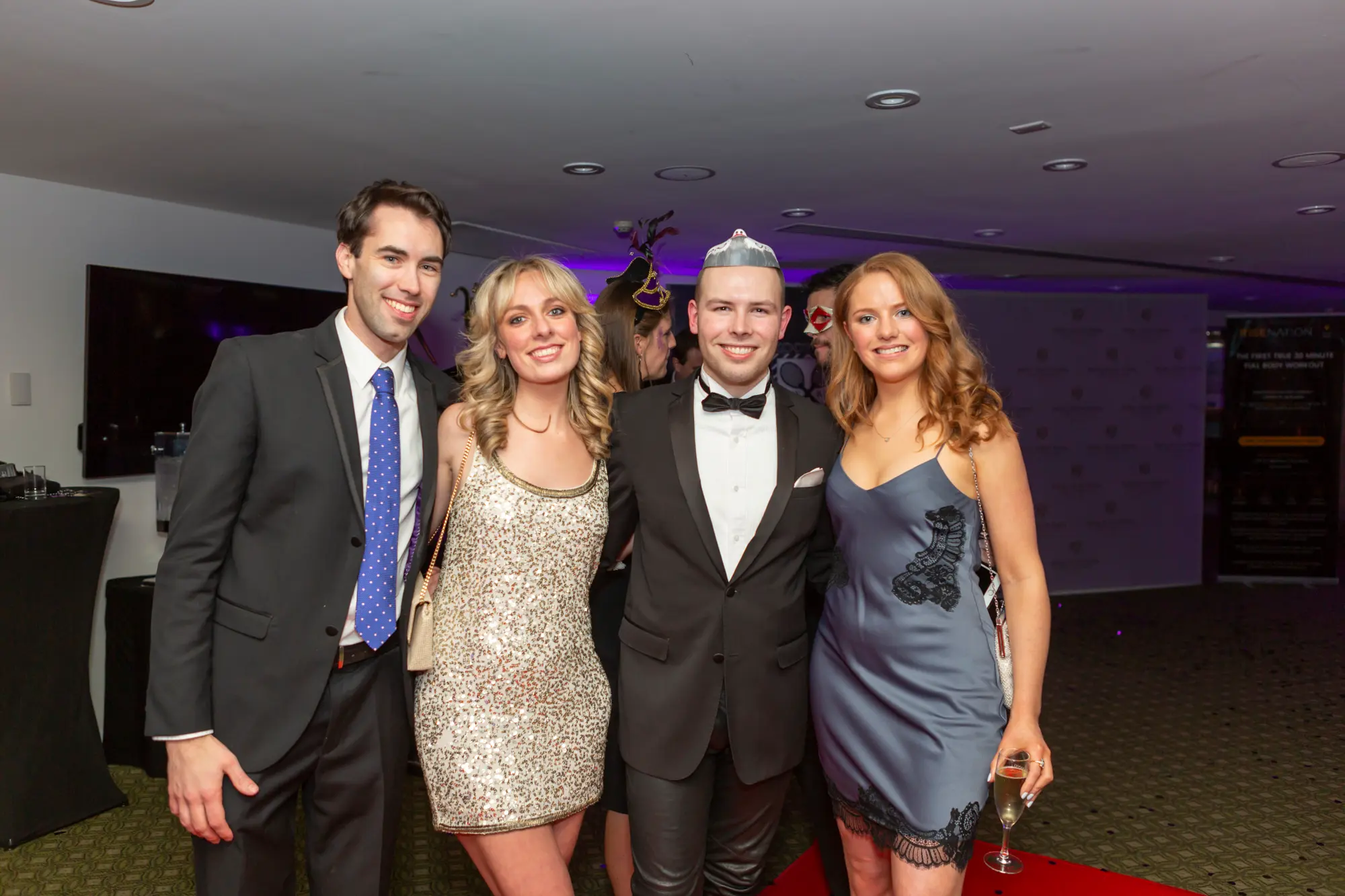 kooyong lawn tennis club masquerade ball yarrayong ball group portrait branded backdrop Four guests pose together at the Kooyong Lawn Tennis Club End-of-Year Masquerade Ball, also known as the YarraYong Ball, held in Toorak. Standing in front of a branded Kooyong and Royal South Yarra backdrop, the group smiles confidently while dressed in formal black-tie attire. Two men wear classic tuxedos, while two women wear elegant evening dresses, with one guest wearing a distinctive silver masquerade-style headpiece. The polished indoor setting and warm lighting reflect the celebratory atmosphere of this annual social event.