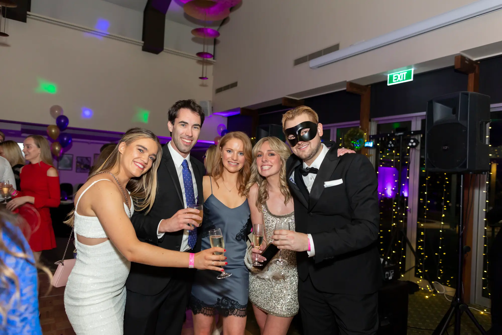 kooyong lawn tennis club masquerade ball yarrayong ball group portrait floral backdrop Five guests pose together during the Kooyong Lawn Tennis Club End-of-Year Masquerade Ball, also known as the YarraYong Ball, held in Toorak. Standing in front of elegant floor-to-ceiling white floral décor with soft purple uplighting, the group smiles and celebrates while dressed in black-tie evening wear. Champagne glasses are raised as part of the festivities, with one guest wearing a classic black masquerade mask. Reflections in the mirrored wall behind them capture the lively atmosphere of this annual social celebration for Kooyong and Royal South Yarra members.