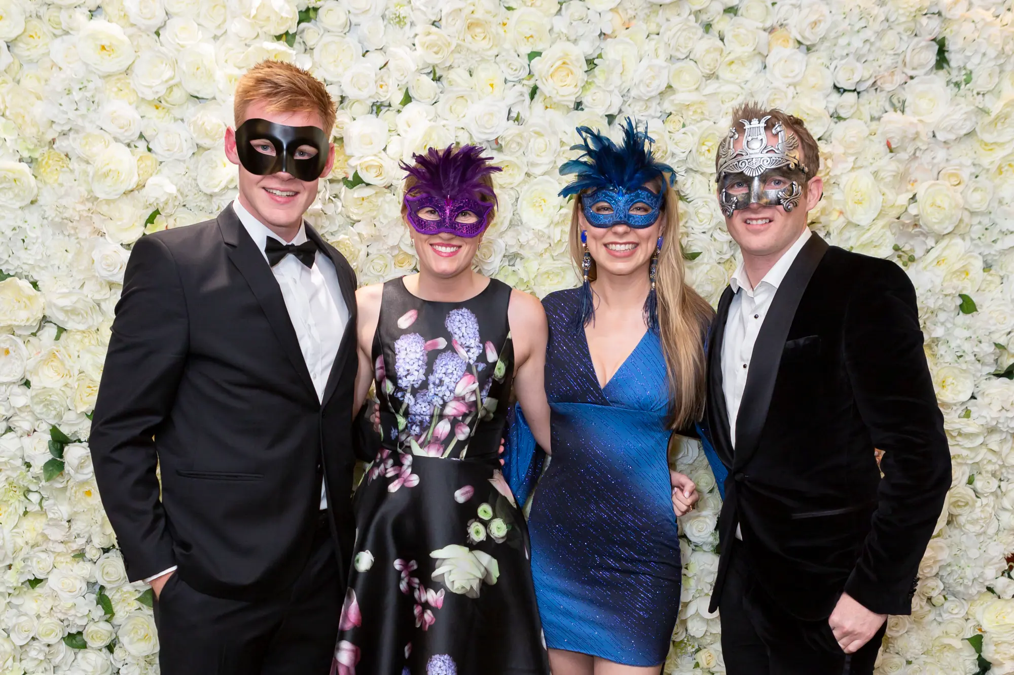 kooyong lawn tennis club masquerade ball yarrayong ball group portrait four guests Four guests pose together for a group photo at the Kooyong Lawn Tennis Club End-of-Year Masquerade Ball, also known as the YarraYong Ball, in Toorak. Two men in black tuxedos and two women in elegant evening dresses stand in front of a white rose floral wall, smiling and wearing colourful masquerade masks. Warm ambient lighting, subtle tennis club memorabilia, and a relaxed celebratory mood highlight this stylish annual event for Kooyong and Royal South Yarra Lawn Tennis Club members.