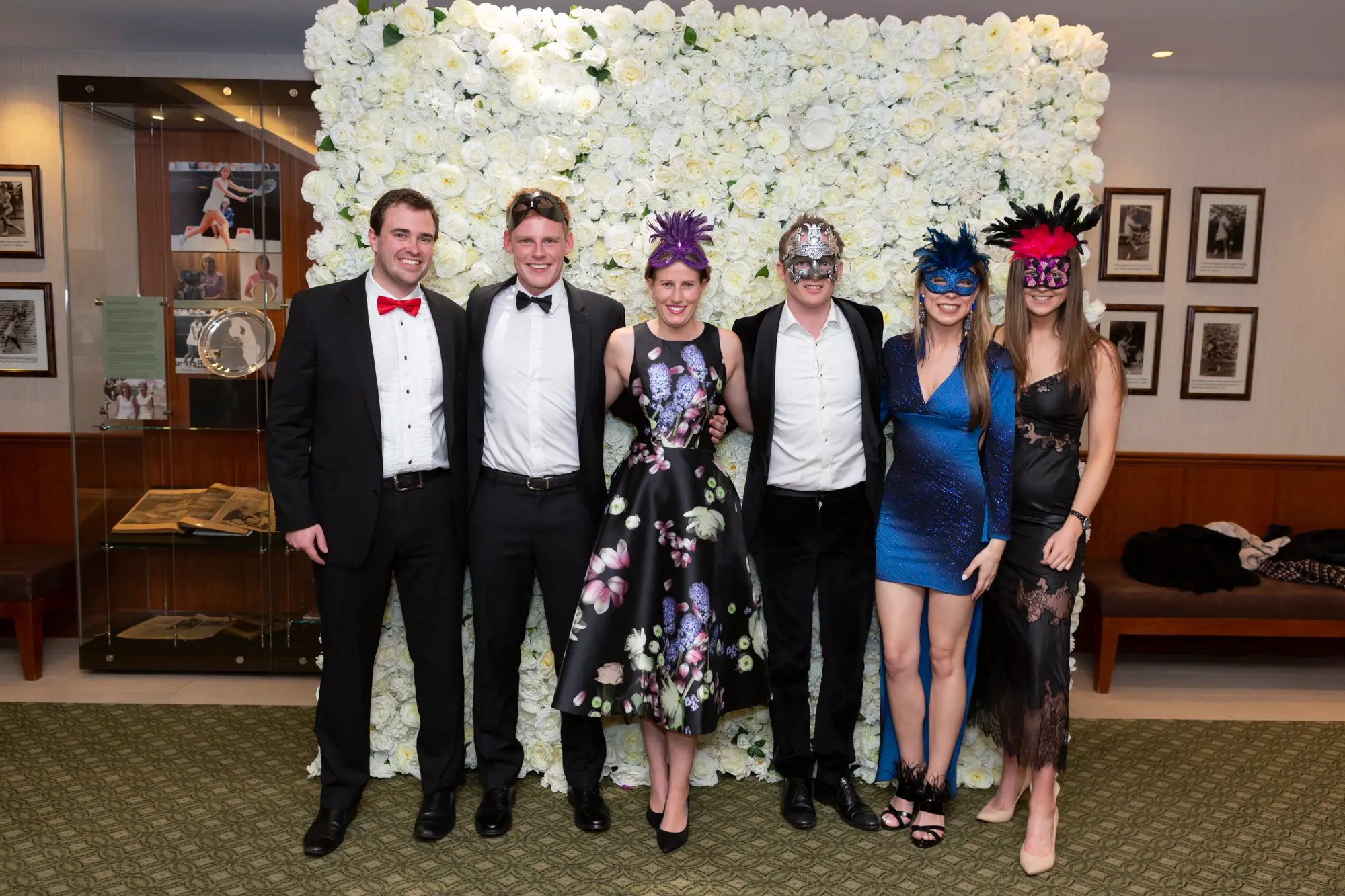 kooyong lawn tennis club masquerade ball yarrayong ball group portrait six guests Six guests pose together for a joyful group portrait at the Kooyong Lawn Tennis Club End-of-Year Masquerade Ball, also known as the YarraYong Ball, held in Toorak. Standing in front of an elegant white rose floral wall, the group smiles warmly while dressed in black-tie evening wear in shades of black, red, green, blue, pink, and gold sequins. Masquerade masks, champagne glasses, warm ambient lighting, and subtle tennis club memorabilia capture the lively, sophisticated atmosphere of this celebrated annual event for Kooyong and Royal South Yarra members.