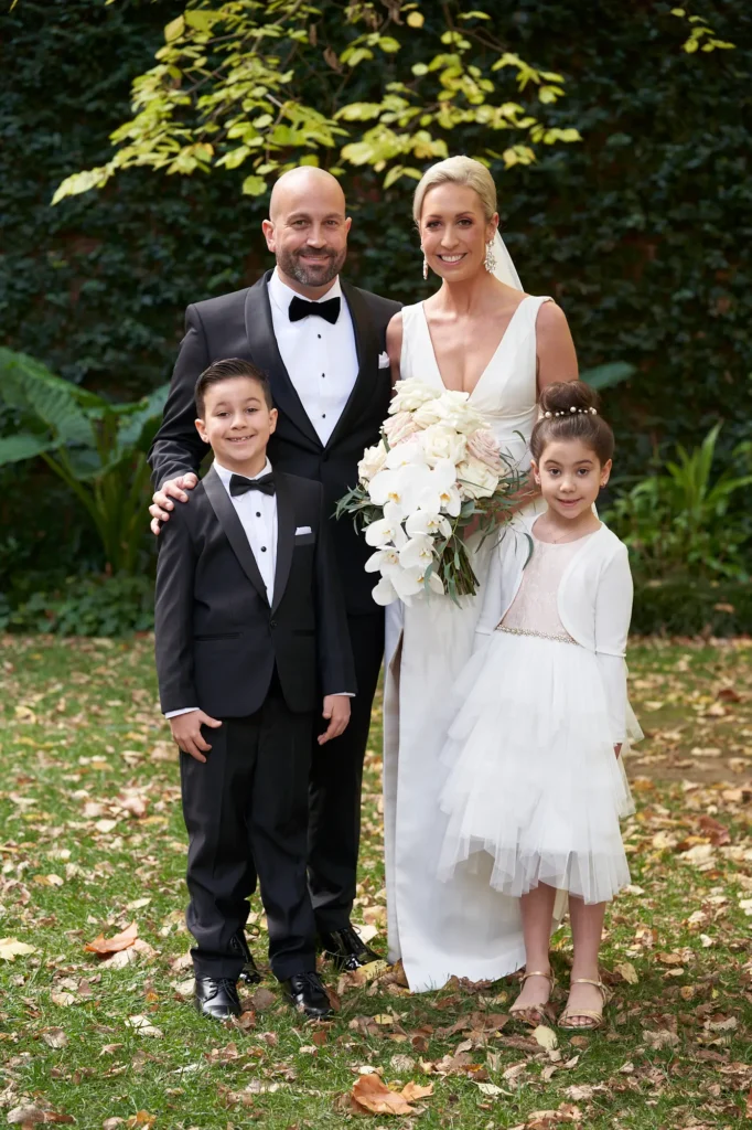 A joyful family portrait of the bride and groom with their children during their wedding day at St Augustine Parish, Melbourne, photographed in a leafy garden courtyard with lush greenery and soft natural light.