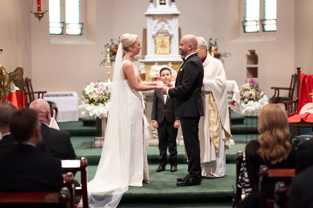 svg%3E Adrian and Laura exchange vows at the altar of St Augustine Parish Church on Bourke Street in Melbourne, standing hand in hand beneath stained glass windows and soft natural light. Surrounded by guests in the pews, the couple share a joyful and intimate moment as the priest presides beside them, with floral arrangements and candles framing the historic church setting.