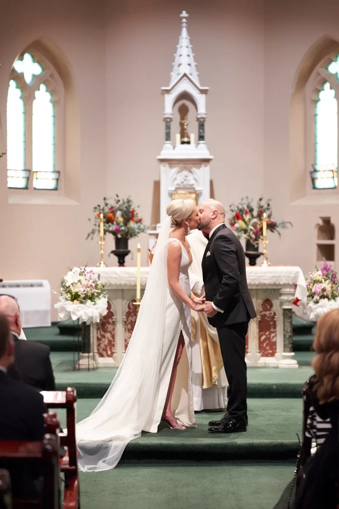 Bride and Groom share their first kiss as a married couple at the altar of St Augustine Parish on Bourke Street, Melbourne, surrounded by floral arrangements and stained glass windows, as the priest looks on and guests watch from the pews during the romantic church wedding ceremony.