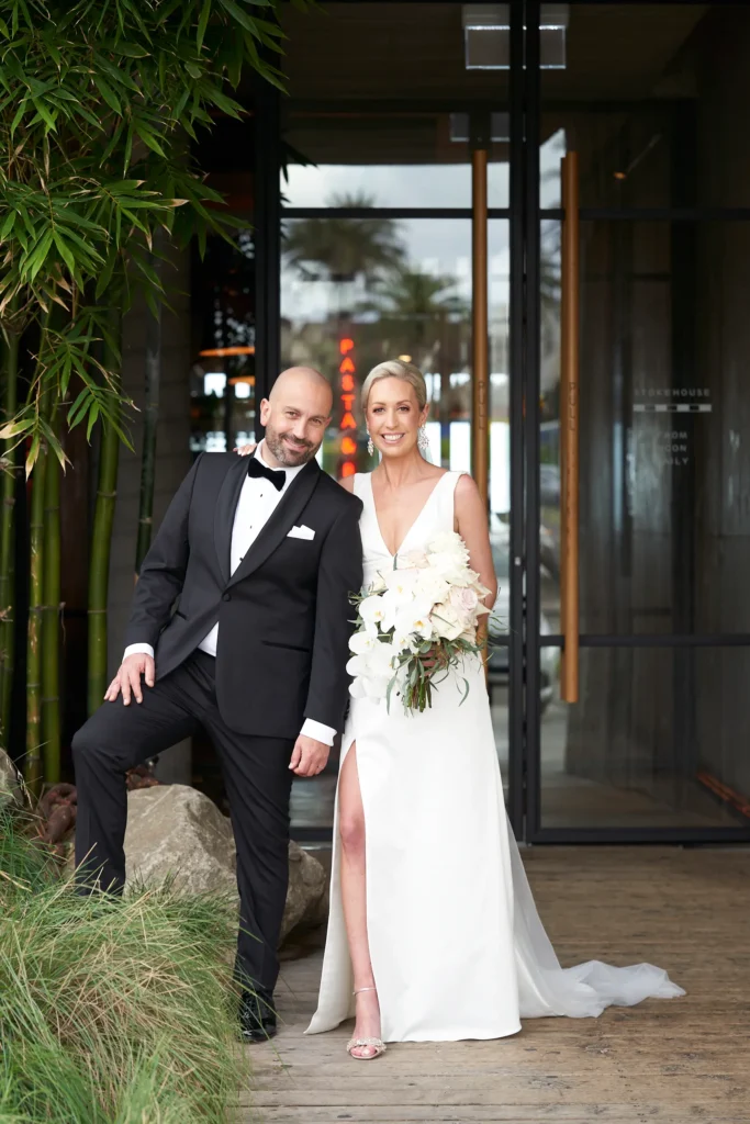 Bride and groom pose together at Stokehouse St Kilda, framed by textured concrete, dark timber slats, and lush bamboo greenery. The groom leans in confidently in a black tuxedo while the bride smiles beside him, holding a cascading orchid bouquet, creating a relaxed and modern wedding portrait filled with effortless elegance.
