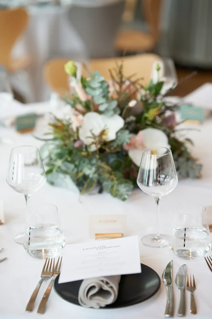 An elegant wedding reception table setting at Stokehouse St Kilda, styled with white linen, black accent plates, grey napkins, printed menus, and crystal glassware. Soft natural light and timber floors create a calm, modern atmosphere, capturing the anticipation and refined detail before guests arrive.