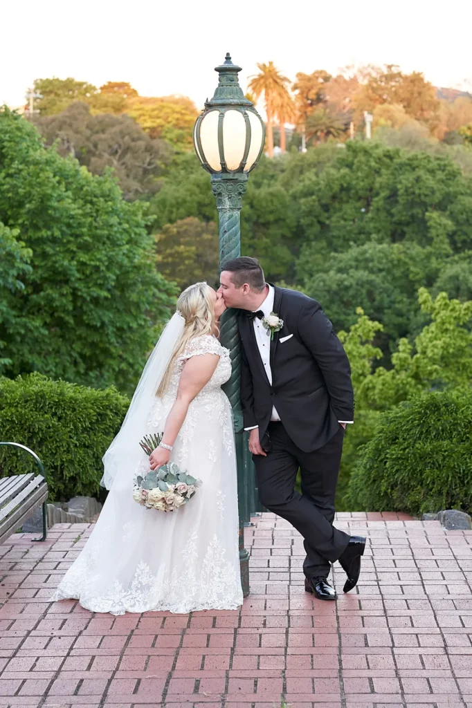 Bride and groom share a romantic kiss beside a vintage green park lamppost at a scenic lookout in O’Connell Reserve opposite Amora Herencia Riverwalk Melbourne. The bride wears a lace A-line wedding gown with veil and holds a bouquet of blush and white roses while the groom in a black tuxedo leans casually against the lamppost overlooking the Yarra River parklands.
