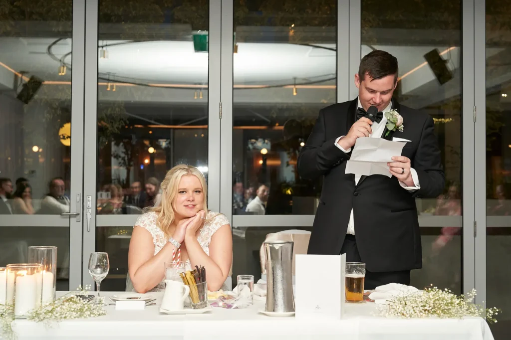 Groom reads from his speech while holding a microphone during the wedding reception at Amora Herencia Riverwalk Melbourne. Standing beside the sweetheart table decorated with baby’s breath flowers and candles, the groom in a black tuxedo speaks to guests as the bride listens beside him, capturing a heartfelt and emotional moment of the Melbourne wedding celebration.