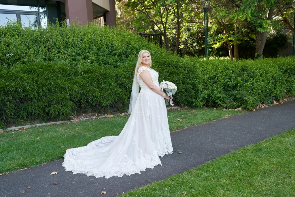 svg%3E Bride smiling radiantly during a bridal portrait in the lush gardens near Amora Hotel Riverwalk Melbourne. She wears a sleeveless lace A-line wedding gown with intricate floral appliqué and a dramatic lace-edged train flowing across the path, with a delicate veil in her hair. Holding a bouquet of white and blush roses with eucalyptus and a trailing ribbon, she turns gracefully toward the camera. A tall evergreen hedge, green lawn, and glimpses of the hotel’s modern windows frame this elegant Melbourne riverside wedding portrait in Burnley Park.