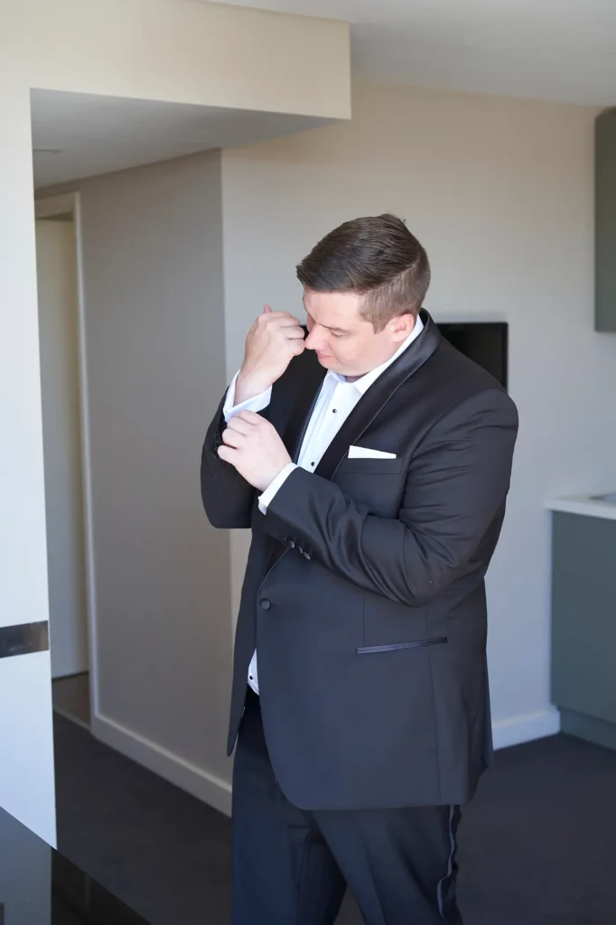 Groom adjusts his cufflinks while getting ready in a bright suite at Amora Herencia Riverwalk Melbourne. Wearing a sharp black tuxedo with satin lapels and a crisp white formal shirt, he looks down with quiet focus as he completes the final touches to his formal look. The modern hotel interior with neutral walls and a sleek kitchenette softly lit by natural daylight creates a calm atmosphere, capturing the groom’s reflective moment before heading to the Melbourne riverside wedding ceremony.