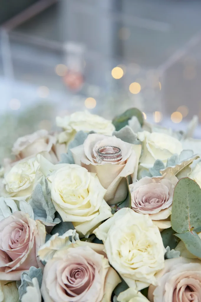 Close-up wedding detail of the bride and groom’s engagement ring and wedding bands resting on a blush rose within the bridal bouquet at Amora Herencia Riverwalk Melbourne. The rings sparkle among soft ivory and dusty pink roses, eucalyptus, and dusty miller leaves, creating an elegant floral setting. A shallow depth of field and warm golden bokeh lights in the background produce a dreamy atmosphere, highlighting the symbolism of the couple’s union during this intimate Melbourne riverside wedding.