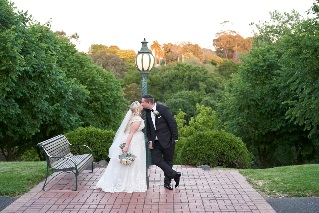 svg%3E Bride and groom share a romantic kiss beside a vintage green lamppost at the scenic Burnley Park lookout near Amora Herencia Riverwalk Melbourne. The bride wears a classic lace A-line wedding gown with a flowing veil and bouquet of roses and greenery, while the groom in a black tuxedo leans casually against the lamppost. Surrounded by manicured hedges, tall trees, and a charming park bench, the red-brick lookout overlooks the peaceful Yarra River valley, where golden hour light creates a warm and timeless Melbourne wedding portrait.