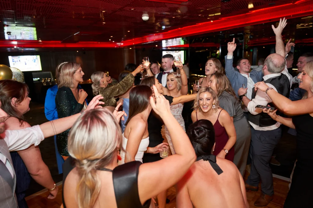 svg%3E Guests dance and raise their glasses in celebration during the Pearses Awards Night & End-of-Year Ball at Eureka 89 Melbourne, capturing the high-energy spirit of the evening. Women in elegant dresses laugh and move across the vibrant dance floor, while others toast in the background beneath a dramatic red-lit ceiling. Surrounded by sleek modern interiors and event screens, the scene reflects the glamour, excitement, and sky-high atmosphere of this prestigious Melbourne awards celebration filled with music, connection, and unforgettable moments.