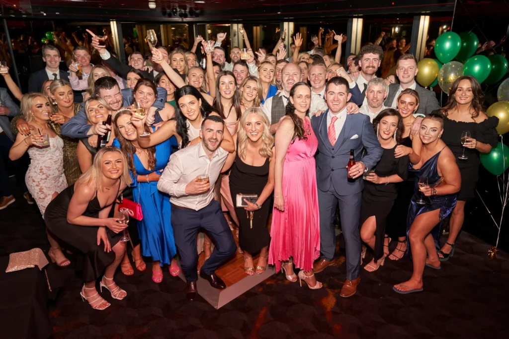 svg%3E A large group of elegantly dressed guests gather on the dance floor raising champagne and beer glasses in a joyful toast during the Pearses Awards Night & End-of-Year Ball at Eureka 89 Melbourne. Smiles, cheers, and raised arms capture the camaraderie and pride of the evening, with bold cocktail dresses and sharp suits reflecting the formal dress code. Framed by green and gold balloons and warm ambient lighting, the modern sky-high venue creates a sophisticated backdrop for this unforgettable Melbourne end-of-year celebration.
