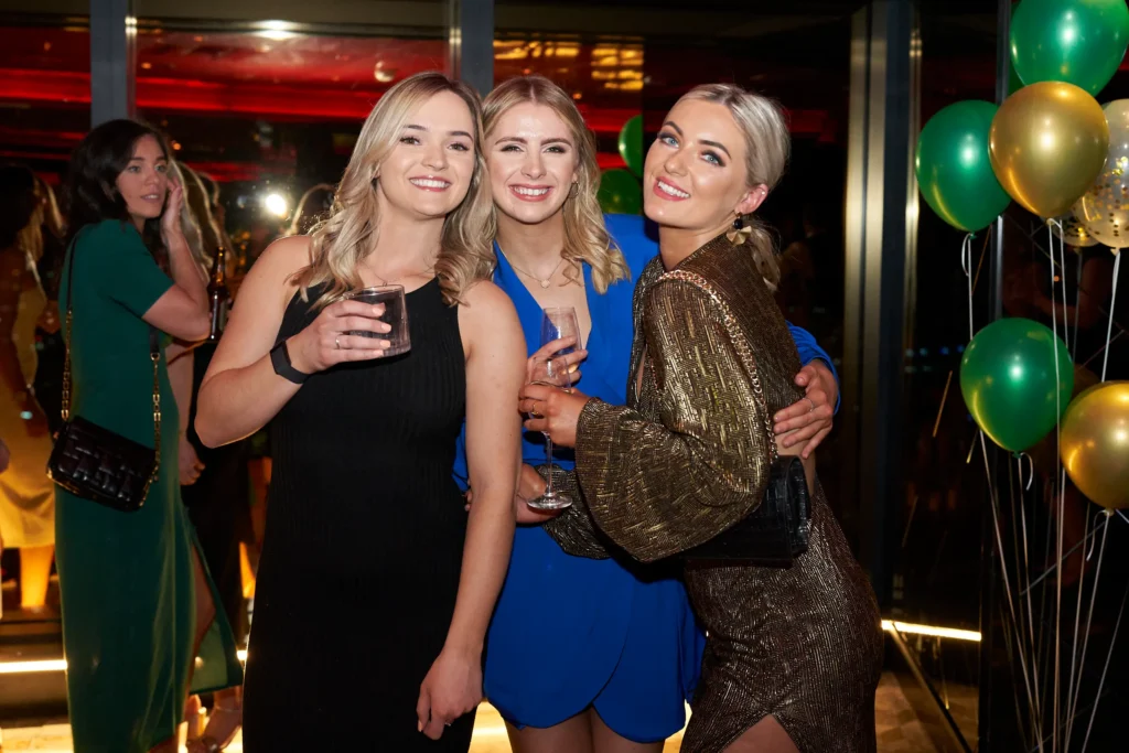 svg%3E Three elegantly dressed women pose arm in arm during the Pearses Awards Night & End-of-Year Ball at Eureka 89 Melbourne, smiling as they hold drinks against a backdrop of green and gold balloons. Dressed in black, vibrant blue, and shimmering gold, they lean together in celebration, capturing the camaraderie and glamour of the evening. Set inside the sky-high venue with floor-to-ceiling windows and formally dressed guests mingling behind them, the image reflects the sophisticated atmosphere, city views, and vibrant energy of this unforgettable Melbourne awards celebration.