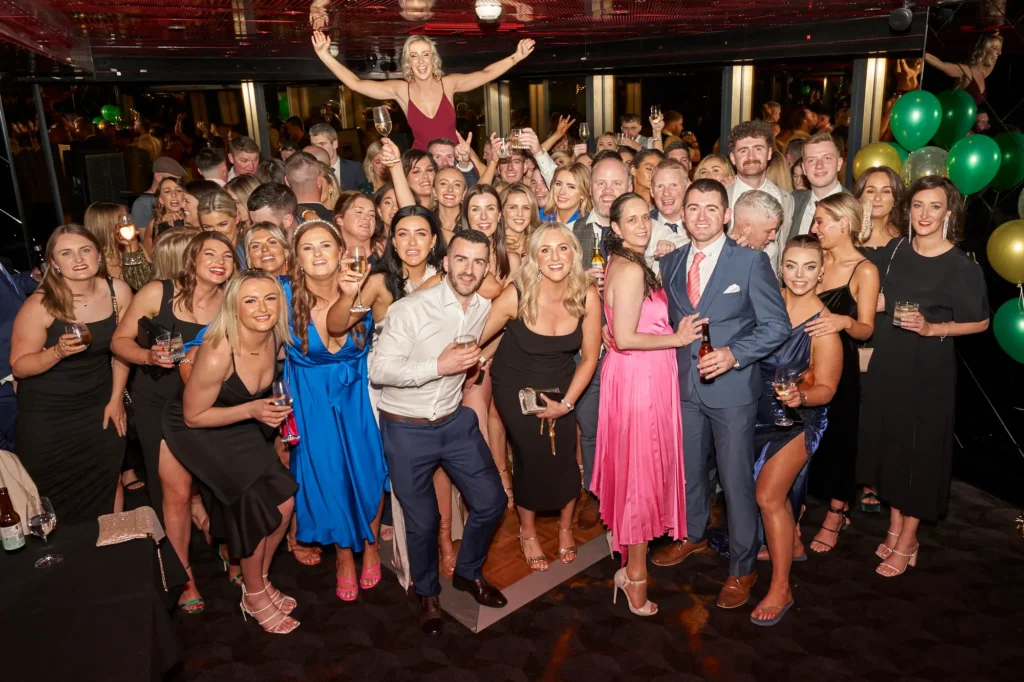 A large group of guests gather for an exuberant group photo at the Pearses Awards Night & End-of-Year Ball at Eureka 89. Dressed in vibrant cocktail dresses and sharp suits, they raise champagne and beer in a collective toast, while one woman is lifted above the crowd with arms outstretched in celebration. Framed by green and gold balloons, a parquet dance floor, and the venue’s signature red-lit ceiling, the image captures a joyful, sky-high end-of-year celebration above Melbourne.