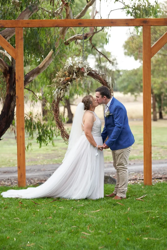 Newlyweds sharing a romantic kiss under rustic timber ceremony arbor at Allurah Functions & Events Ballarat, framed by native eucalyptus wreath and countryside landscape, bride in lace gown and groom in blue blazer.