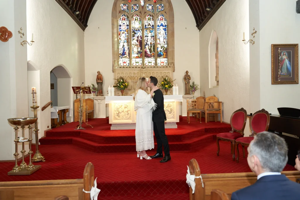 svg%3E Bride and groom share their first kiss as husband and wife at St John Catholic Church in Heidelberg, Melbourne, framed by a vibrant multi-panel stained glass window above the altar. The bride wears a long-sleeved white lace gown and block-heeled sandals while the groom leans in wearing a black suit. Guests sit in dark wooden pews, surrounded by floral arrangements and traditional church details, capturing an intimate, solemn, and romantic wedding moment full of warmth and timeless elegance.