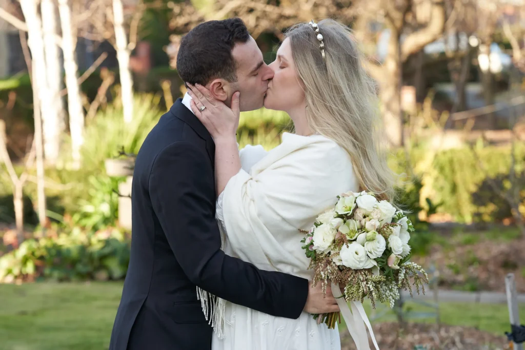 svg%3E Bride and groom share a tender kiss in a secluded garden on the grounds of St John Catholic Church in Heidelberg, Melbourne. The groom, in a black tuxedo, gently cups the bride’s face while she rests her hand on his neck, holding a bouquet of white roses and seasonal greenery. Surrounded by lush greenery, soft lawn, bare winter-spring trees, and a stone birdbath, the couple is captured in an intimate, peaceful, and romantic portrait with soft natural light highlighting the elegance of her beaded headband and cream pashmina wrap.