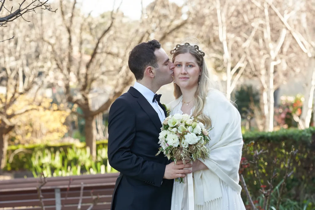 svg%3E Bride and Groom share a tender wedding moment in a lush garden near St John Catholic Church in Heidelberg, Melbourne. The groom, in a black tuxedo, gently kisses the bride on her cheek as she faces the camera wearing a white wedding dress and cream-colored shawl. She holds a hand-tied bouquet of white roses, carnations, lisianthus, and greenery, while wearing an ornate circular headband. Surrounded by manicured hedges, soft foliage, and bare winter-spring trees, the couple is captured in a peaceful, intimate, and timelessly elegant outdoor wedding portrait.
