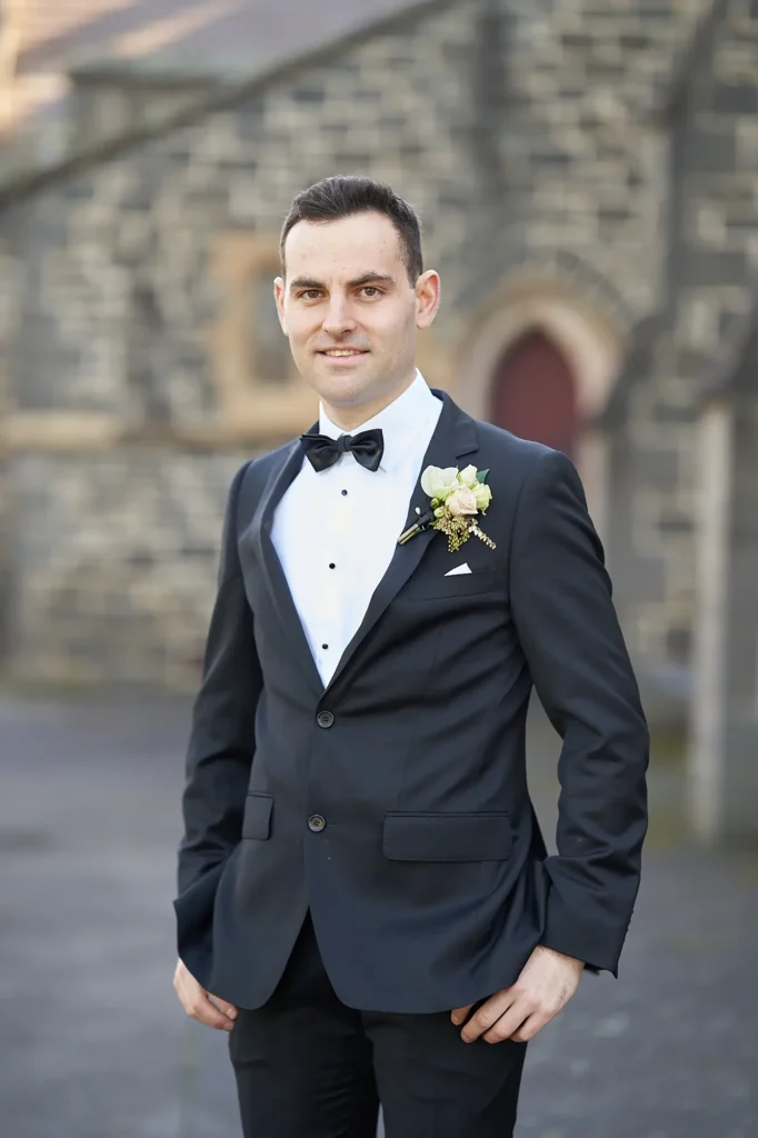 Groom stands confidently on the grounds of St John Catholic Church in Heidelberg, Melbourne, wearing a black tuxedo, white dress shirt, and black bow tie. His hands are casually tucked in his trouser pockets, complemented by a white pocket square and boutonniere of white roses with dried foliage. The historic bluestone church and an arched red doorway provide a soft-focus backdrop, creating a timeless, elegant portrait that captures the groom’s calm, polished presence and quiet anticipation on his wedding day.