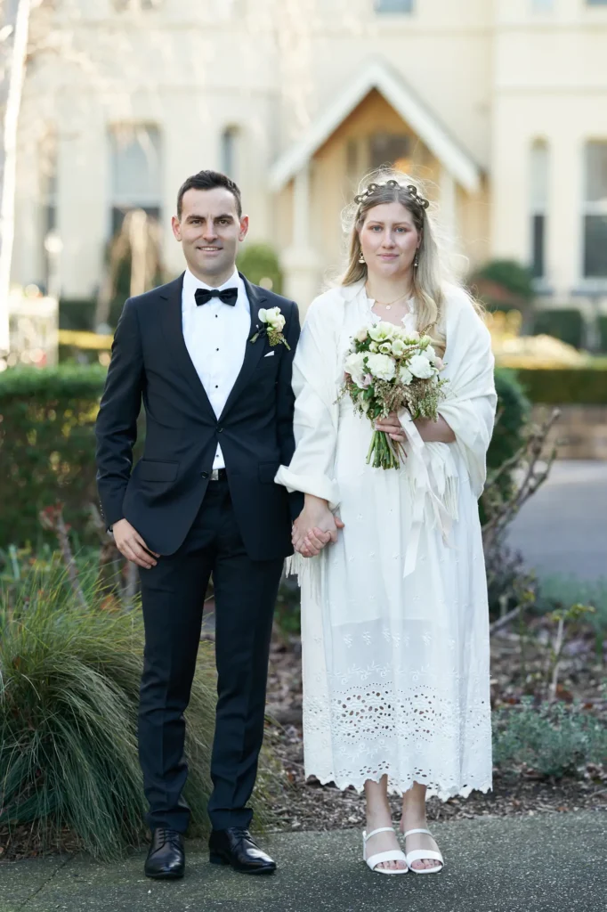 Formal wedding portrait of the bride and groom on a paved garden path at St John’s Catholic Church, Heidelberg. The groom wears a black tuxedo with a black bow tie and white rose boutonniere. The bride wears a long-sleeved white lace gown with an eyelet-embroidered hem, a cream-colored fringe pashmina, white block-heeled sandals, and a gold circular headband. They hold hands and face the camera, framed by manicured green hedges, dormant garden beds, slender bare trees, and the cream-colored parish presbytery, creating a composed, timeless, and serene atmosphere.