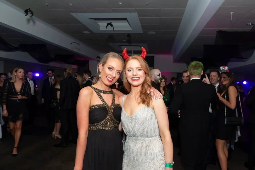 svg%3E Two women pose together at the Kooyong Tennis Club Halloween Gala, one in an elegant black gown with gold beaded embroidery and the other in a shimmering silver sequined dress with glittery red devil horns, while guests in colorful costumes mingle in the background, under sheer black fabric and bat decorations, capturing the luxurious yet playful and spooky atmosphere of this high-energy end-of-year ball in Melbourne.