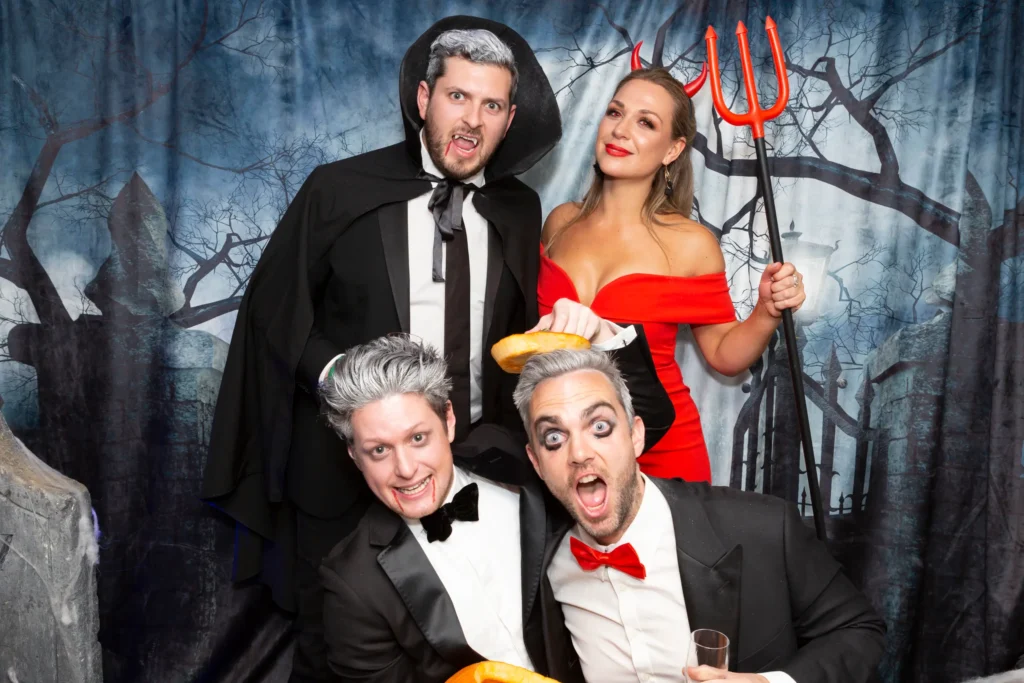 svg%3E A theatrical group at the Kooyong Tennis Club Halloween Gala features a man in a vampire cape with bared fangs, another with spiked grey hair and a velvet bowtie, a woman in a red off-shoulder dress holding a pitchfork with matching devil horns, and two ghoulish men with grey-toned hair and red bowties playfully holding glazed donuts over each other’s heads, posed in front of a cemetery-themed backdrop with iron gates, tombstones, and twisted trees, capturing the lively, spooky, and luxurious end-of-year ball atmosphere in Melbourne.