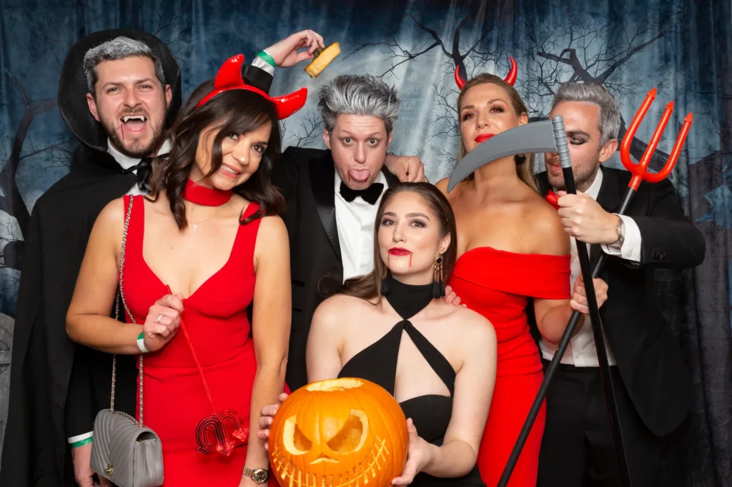A lively group of seven poses for a theatrical portrait at the Kooyong Tennis Club Halloween Gala, Royal South Yarra. Three men in vampire-inspired makeup and grey-toned hair stand behind two women in red cocktail dresses with glossy devil horns, holding a trident and tail prop. In the foreground, a woman in a black halter-neck dress holds a carved jack-o'-lantern with a stitched-mouth expression. They are positioned in front of a cemetery-themed backdrop with twisted trees and stone pillars. Crisp professional lighting highlights their formalwear, elaborate makeup, and the sophisticated “Haunted Gala” atmosphere.