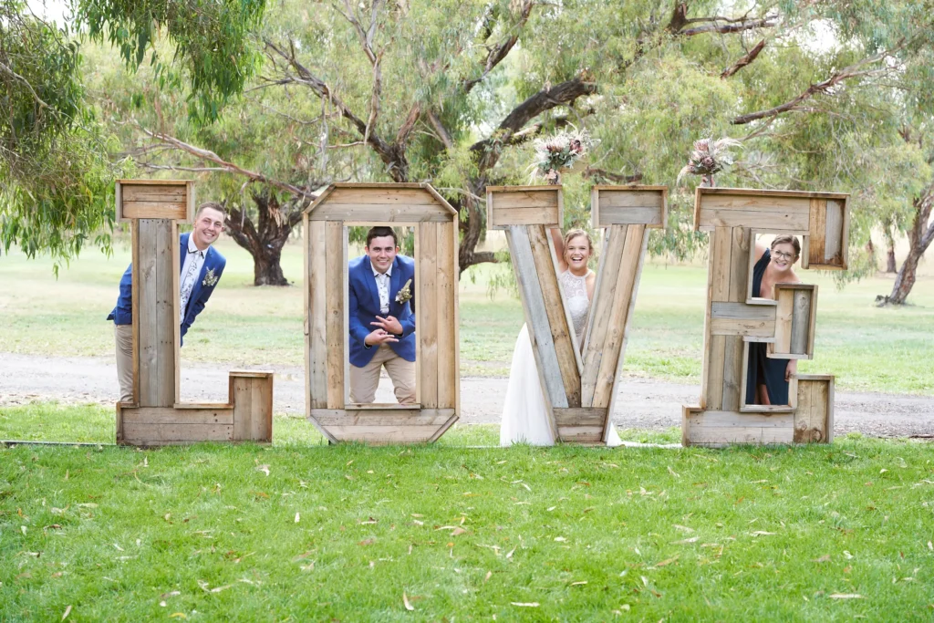 svg%3E Playful bridal party posing with rustic LOVE letters at Allurah Functions & Events in Ballarat, featuring bride Miriam smiling behind the V with bouquet, groomsmen in blue suits peeking through L and O, and bridesmaid in navy beside E, surrounded by eucalyptus trees and lush countryside lawn, capturing a joyful and relaxed Australian country wedding atmosphere.
