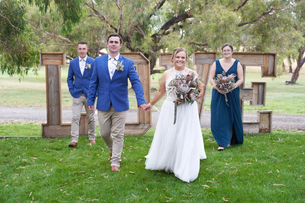 svg%3E Bride and groom walking hand in hand across a grassy lawn at Allurah Functions & Events in Ballarat, with bridal party following behind and rustic LOVE letters in the background, bride holding a native protea bouquet and groom in a blue blazer, surrounded by eucalyptus trees in a relaxed and romantic countryside wedding setting.