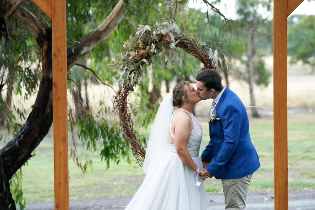 svg%3E Bride and groom sharing their first kiss during an outdoor wedding ceremony at Allurah Functions & Events in Ballarat, standing beneath a rustic circular floral arch with eucalyptus and native foliage, holding hands as the bride’s veil flows softly, framed by natural countryside scenery and gum trees, capturing a romantic and intimate moment in a serene country wedding setting.