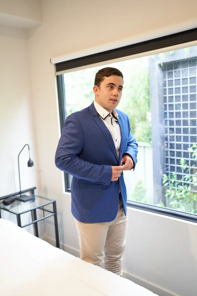 Groom completing his wedding preparation by buttoning a blue suit jacket in a minimalist bedroom in Ballarat, shown in a calm three-quarter profile with a contemplative expression, wearing a white dress shirt with black accents and tan chinos, standing in soft natural window light with blurred garden greenery outside, capturing a quiet and elegant final getting-ready moment before the countryside ceremony.