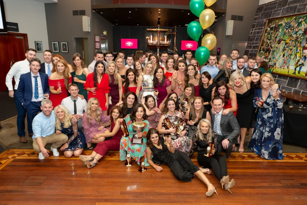 svg%3E A large group of nearly 50 attendees gather on the dance floor at the Padraig Pearses GAA Club End of Year Gala Dinner & Awards at Carlton Brewhouse Abbotsford. Guests are arranged standing, kneeling, and sitting on the polished wooden floor, with several front-center individuals holding gold and silver trophies. Behind them, polished copper brewery tanks, glowing digital screens, a GAA match painting, and green and gold balloons set the celebratory scene, capturing the lively, triumphant, and joyful atmosphere of the club’s milestone awards night in Melbourne.