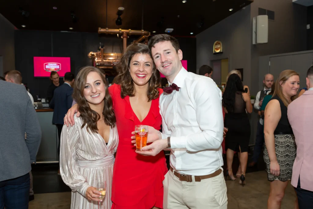 svg%3E Three attendees pose together at the Padraig Pearses GAA Club End of Year Gala Dinner & Awards at Carlton Brewhouse Abbotsford. The central woman wears a vibrant red ruffled dress holding a glass of Carlton Draught beer, flanked by a woman in a champagne pleated dress with sparkling wine and a man in a white dress shirt with a dark bow tie. Behind them, a polished copper beer tank and glowing Carlton Brewhouse signage highlight the industrial-chic bar setting, while blurred guests in formal attire create a lively, celebratory atmosphere.