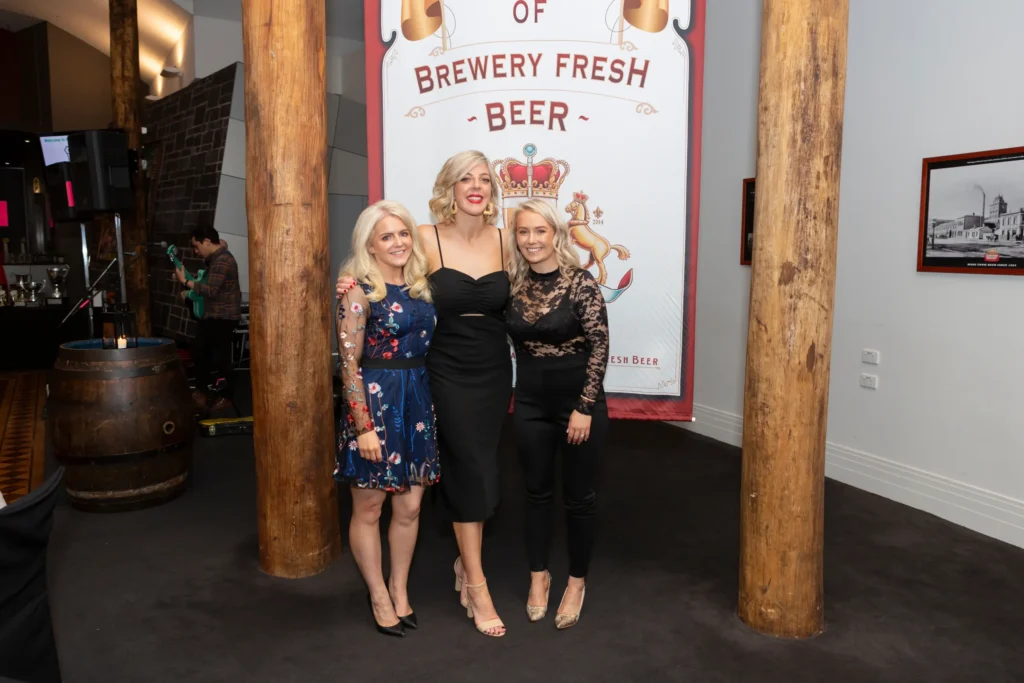 svg%3E Three women pose together at the Padraig Pearses GAA Club End of Year Gala Dinner & Awards at Carlton Brewhouse Abbotsford, smiling warmly in cocktail attire. The central woman wears a sleek black midi dress, flanked by friends in a floral blue dress and black lace top with trousers. Behind them, rustic brewery décor includes a large banner reading "BREWERY FRESH - BEER -", wooden pillars, a beer barrel, and a musician playing guitar, capturing the lively, celebratory atmosphere of the club’s intimate and stylish awards night in Melbourne.