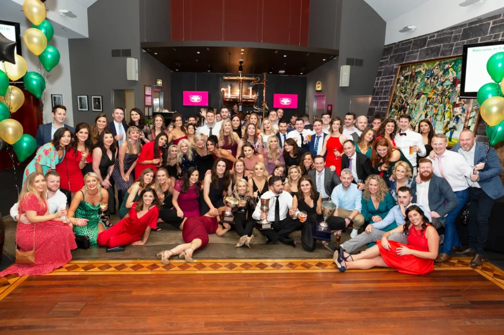 Large group celebrating Padraig Pearses GAA Club End of Year Gala Dinner & Awards at Carlton Brewhouse Abbotsford, posing in a victory group shot with silver trophies, cheering with raised drinks and smiling together on the dance floor, capturing a triumphant club celebration and season success.