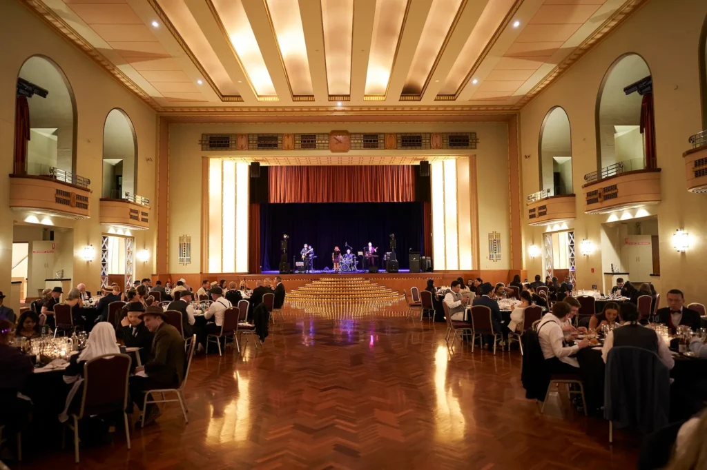 Wide view of a grand ballroom during a Melbourne awards night and gala dinner, with banquet tables, guests, a live band, and an elegant stage.