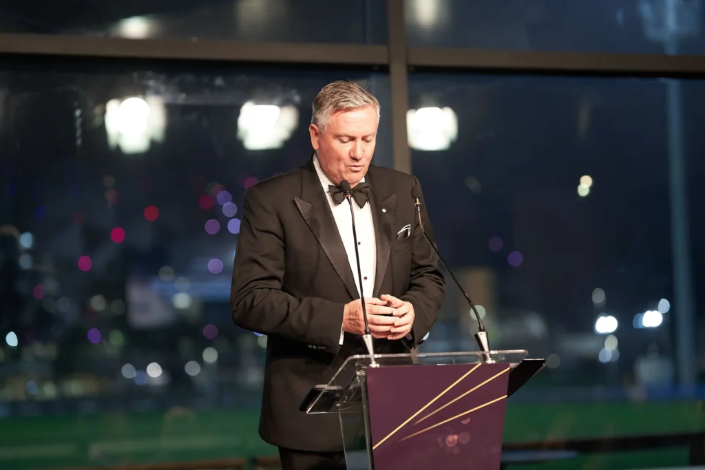 svg%3E Distinguished man in a black tuxedo speaks at a transparent lectern during a prestigious corporate awards gala, with microphones, warm stage lighting and a blurred Melbourne cityscape glowing through the windows behind him.