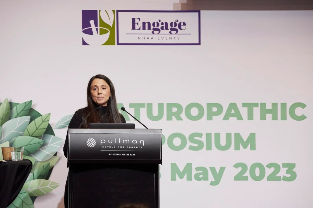 svg%3E Professional speaker addresses an audience from a podium at a naturopathic symposium in Melbourne, with a branded conference backdrop, botanical graphics and Pullman Melbourne Albert Park signage creating a polished corporate event setting.