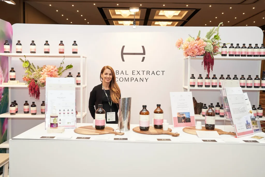 svg%3E Smiling brand representative welcomes attendees from a polished wellness exhibition booth, surrounded by branded product displays, amber bottles, pink roses and elegant floral arrangements at a professional corporate event in Melbourne.