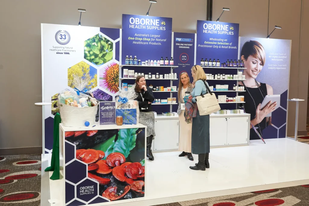 Three women engage in a professional conversation at a medical trade show exhibition booth in Melbourne, surrounded by branded healthcare products, display shelving and promotional materials, capturing an authentic moment of networking and product consultation at a corporate event.