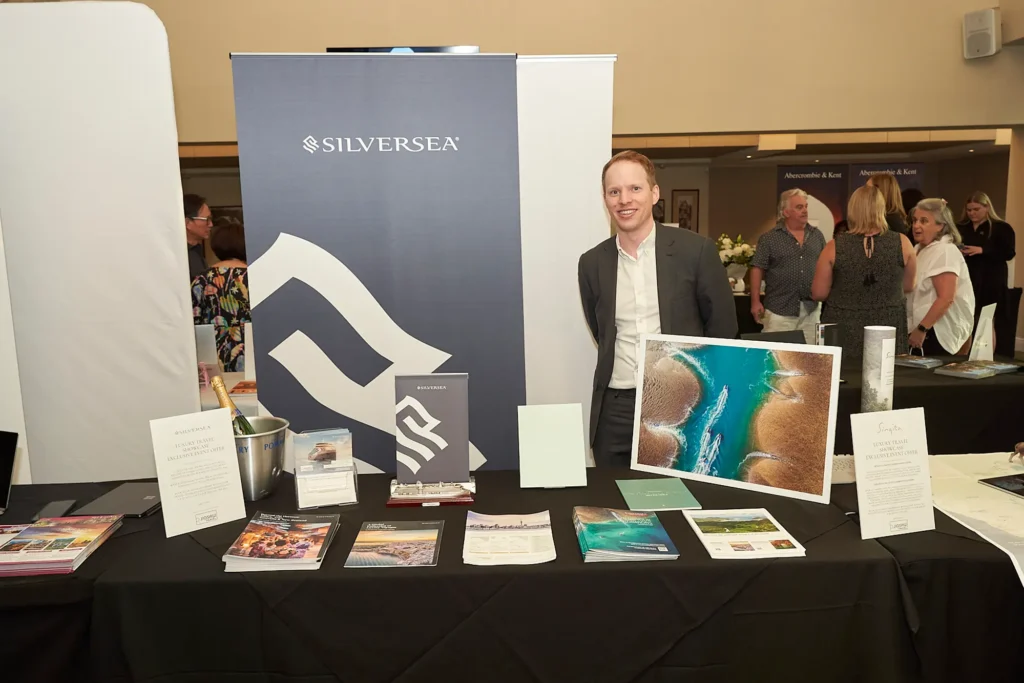 Smiling travel brand representative welcomes visitors at a luxury travel exhibition in Melbourne, standing beside a SilverSea display with cruise brochures, a miniature ship, promotional materials and an ocean landscape photograph in a professional convention setting.