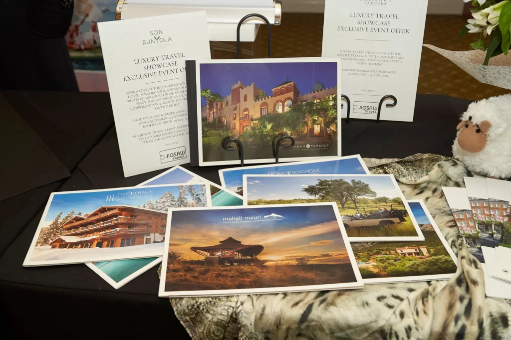 Luxury travel exhibition display featuring glossy destination brochures, promotional cards and exclusive event offers for luxury properties, arranged on a presentation table with a decorative white sheep and patterned fabric at a Melbourne trade exhibition.