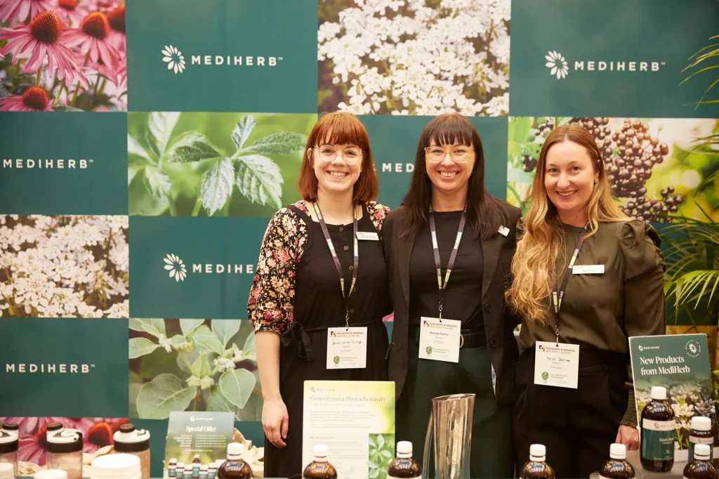 Three smiling brand representatives pose together behind a MediHerb exhibition counter in Melbourne, surrounded by herbal supplement products, botanical graphics and branded materials, capturing a welcoming corporate team moment at a professional trade show.